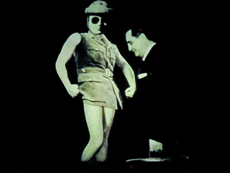 A black-and-white film still showing a female figure in a hat and military-style clothing standing against a dark background, with a second figure partially visible beside her.