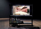 Yuri Pattison, "the ideal (v/0.3.2)", 2015–2021. Vidéo couleur, son. Rayonnage, moniteur, lecteur multimédia, BITMAIN Antminer Cl, unité de refroidissement par eau pour PC, réservoir (Mountain Storm/RW Keeton), pierres de la rivière Dadu, miniatures à l'échelle 1/12, cadenas BROCKHAGE®, autocollant, câbles, cartons d’emballage, eau purifiée, poussière. 30 min 2 sec. Collection Mudam Luxembourg. Acquisition 2022. Vue de l’exposition "Post-Capital : Art et économie à l'ère du digital", Mudam Luxembourg, 02.102021 – 16.01.2022 © Photo : Rémi Villaggi | Mudam Luxembourg