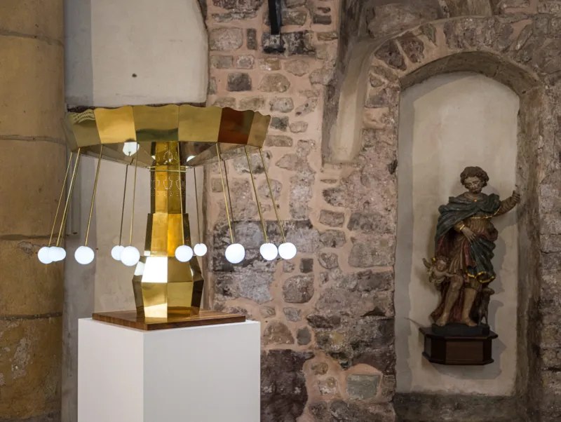 An installation in a historic church interior. An automated miniature carousel of white lights stands beside a Catholic religious figure.