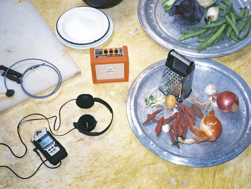 A wooden table with a recording device, a small speaker, old Walkman headphones, two metal plates of vegetables and a grater.