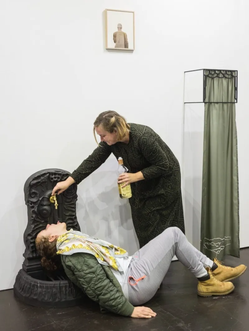 A woman holding a bottle of limoncello turns on a tap on an artistic fountain, pouring a drink for a man lying underneath the spout.