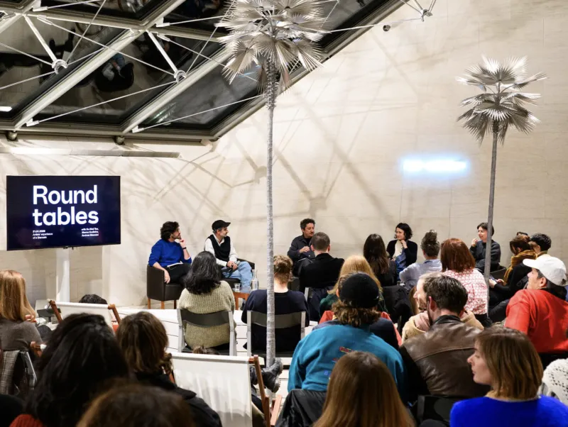 People seated in a Mudam gallery facing a TV screen and a panel of people giving a lecture.
