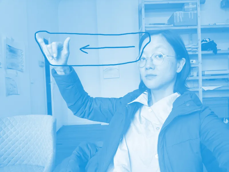 Une femme portant des lunettes et une veste sombre lève la main pour effectuer un geste en langue des signes. Une flèche bleue dessinée à la main pointe vers la gauche à travers l’image.