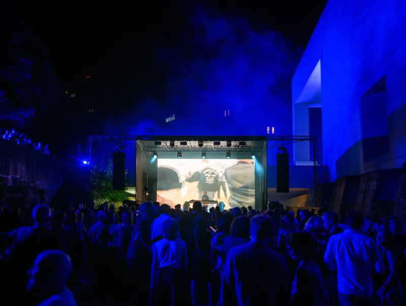 A DJ performs on a large stage at night, with a large screen behind him showing people dancing. A crowd dances in front of the stage near Mudam.