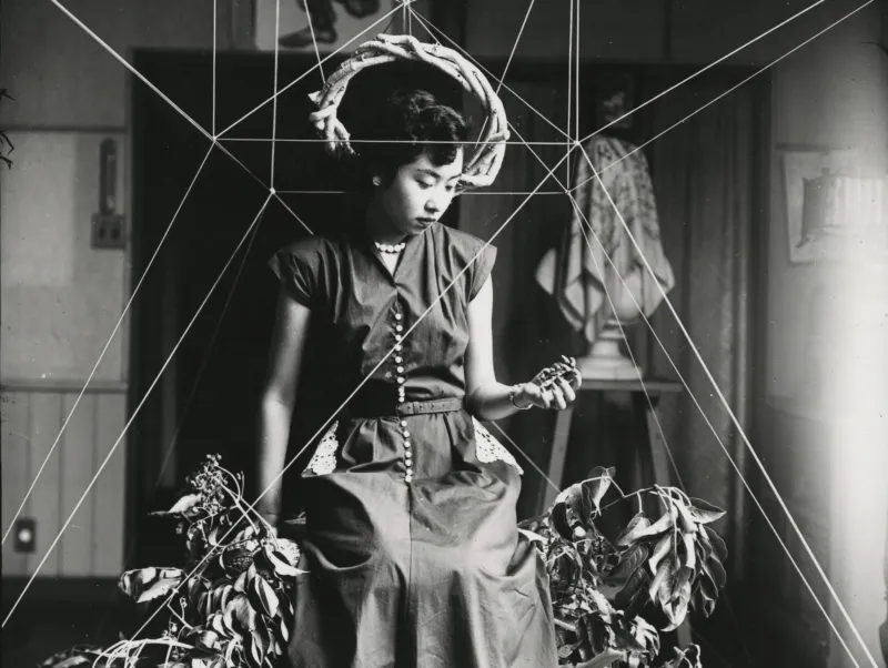 Black-and-white photo of a Japanese woman sitting on a floral structure suspended from the ceiling by thin white strings.