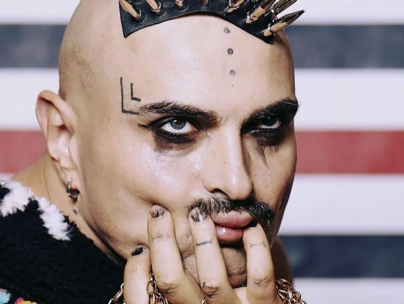 A tattooed man with a mustache and makeup, wearing a spiked silver headpiece, looks boredly upward.