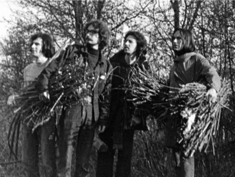 A black-and-white photo of four people standing in front of a wooded area, holding branches in their hands.