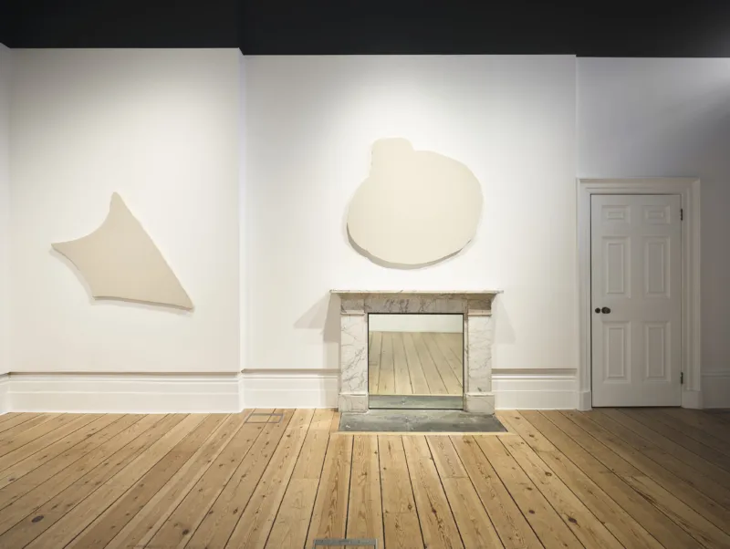 Empty gallery room with wooden floors, a fireplace and two abstract wall sculptures.