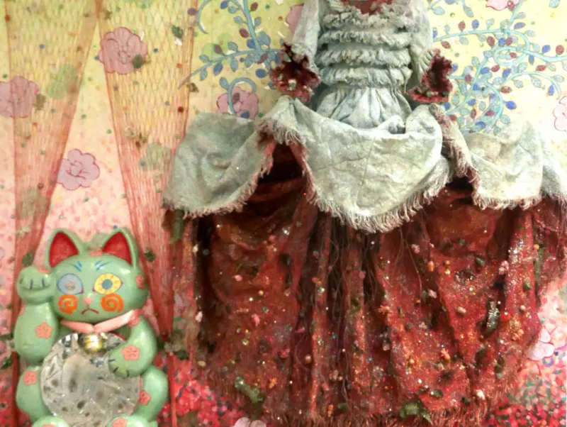 Close-up view of a colorful textile art installation featuring red, patterned fabric, as well as a small green lucky cat figurine, set against a pastel-colored, floral-patterned background.