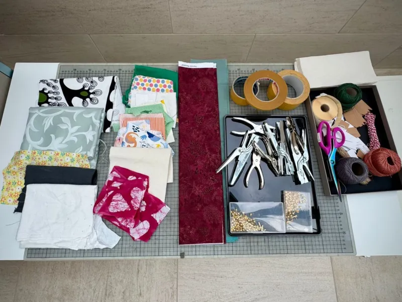 Un espace de couture et de loisirs créatifs avec divers morceaux de tissu colorés, des ciseaux, des perforatrices, des rouleaux de ruban adhésif, des bobines de fil et d’autres fournitures, disposés sur un tapis de découpe quadrillé.