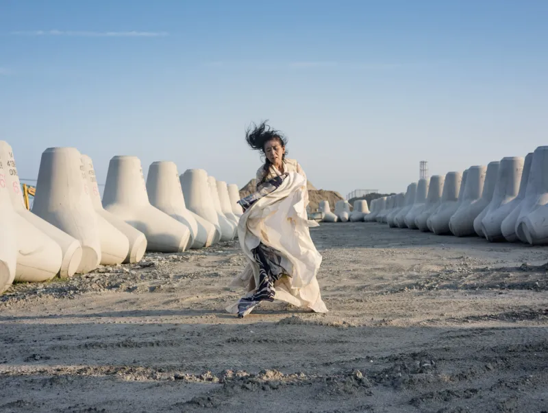 Une femme vêtue de blanc et de vêtements fluides se tient dans un paysage côtier marqué par le béton, entourée de rangées de grands brise-lames, sous un ciel bleu dégagé.