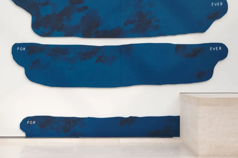 Ein minimalistischer Galerieraum mit einer großen, abstrakten blauen Wandskulptur aus unregelmäßigen horizontalen Formen. Sie ist an einer weißen Wand über einem hellen Steinboden angebracht. Rechts sind eine niedrige weiße Sitzbank und ein Fenster zu sehen.