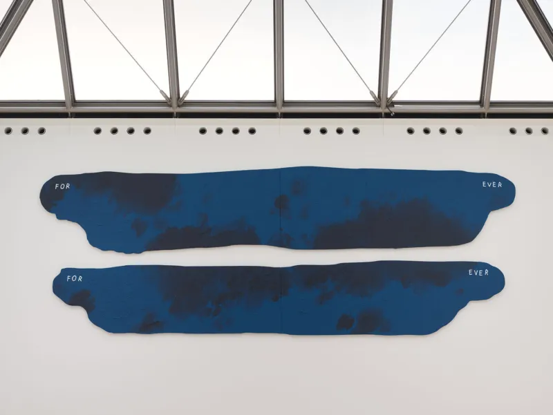 Ein minimalistischer Galerieraum mit einer großen, abstrakten blauen Wandskulptur aus unregelmäßigen horizontalen Formen. Sie ist an einer weißen Wand über einem hellen Steinboden angebracht. Rechts sind eine niedrige weiße Sitzbank und ein Fenster zu sehen.