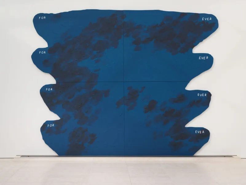Ein minimalistischer Galerieraum mit einer großen, abstrakten blauen Wandskulptur aus unregelmäßigen horizontalen Formen. Sie ist an einer weißen Wand über einem hellen Steinboden angebracht. Rechts sind eine niedrige weiße Sitzbank und ein Fenster zu sehen.