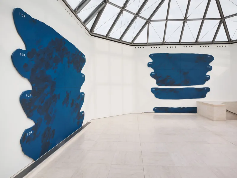 Ein minimalistischer Galerieraum mit einer großen, abstrakten blauen Wandskulptur aus unregelmäßigen horizontalen Formen. Sie ist an einer weißen Wand über einem hellen Steinboden angebracht. Rechts sind eine niedrige weiße Sitzbank und ein Fenster zu sehen.