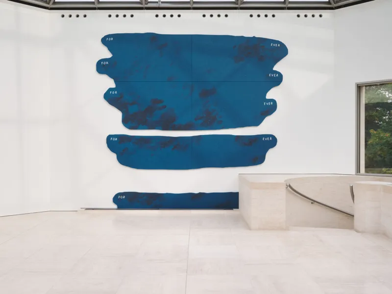 Ein minimalistischer Galerieraum mit einer großen, abstrakten blauen Wandskulptur aus unregelmäßigen horizontalen Formen. Sie ist an einer weißen Wand über einem hellen Steinboden angebracht. Rechts sind eine niedrige weiße Sitzbank und ein Fenster zu sehen.
