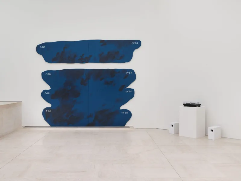 Ein minimalistischer Galerieraum mit einer großen, abstrakten blauen Wandskulptur aus unregelmäßigen horizontalen Formen. Sie ist an einer weißen Wand über einem hellen Steinboden angebracht. Rechts sind eine niedrige weiße Sitzbank und ein Fenster zu sehen.