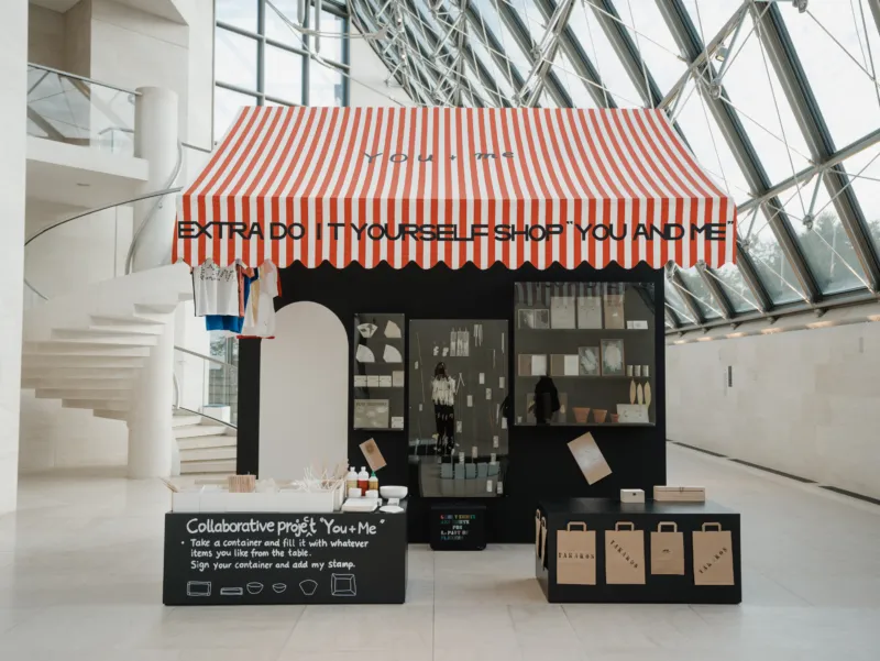 Vue de l’installation de Extra Do It Yourself Shop « You and Me » (1994/2021) de Takako Saito, avec son auvent à rayures rouges et blanches, ses étagères présentant des objets et des matériaux, ainsi qu’un comptoir dédié à un projet collaboratif, au Mudam Luxembourg.