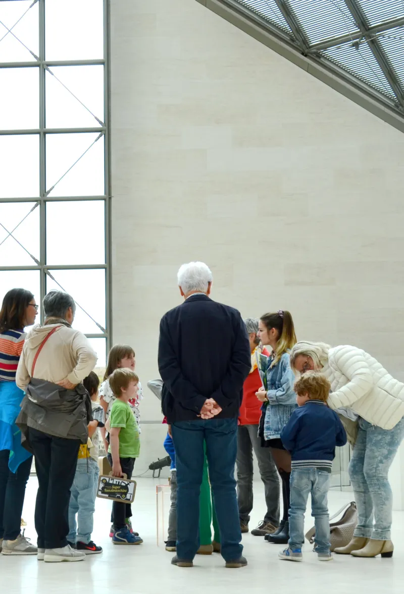 Des adultes et des enfants se rassemblent autour d’une petite installation au sol dans une galerie lumineuse, se penchant avec curiosité et concentration silencieuse.