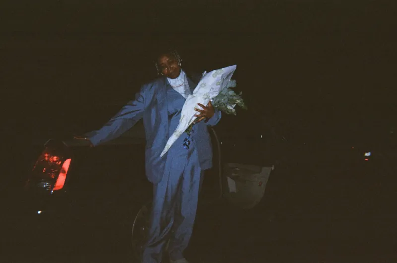 A person holding a flower bouquet while leaning on the back of a limousine.