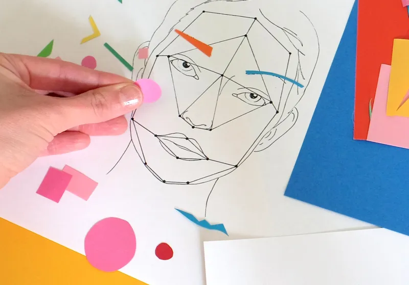 A hand placing papers of different colours, shapes and sizes on the drawing of a face.