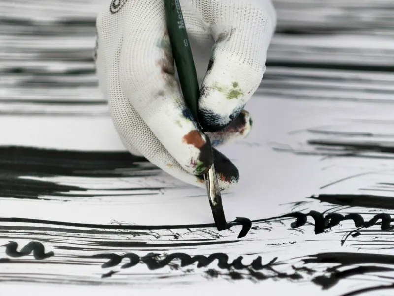 Focus on a white gloved hand painting in black with a brish on a white paper.
