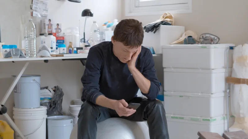 A masked man sitting in a chair checking his smartphone.