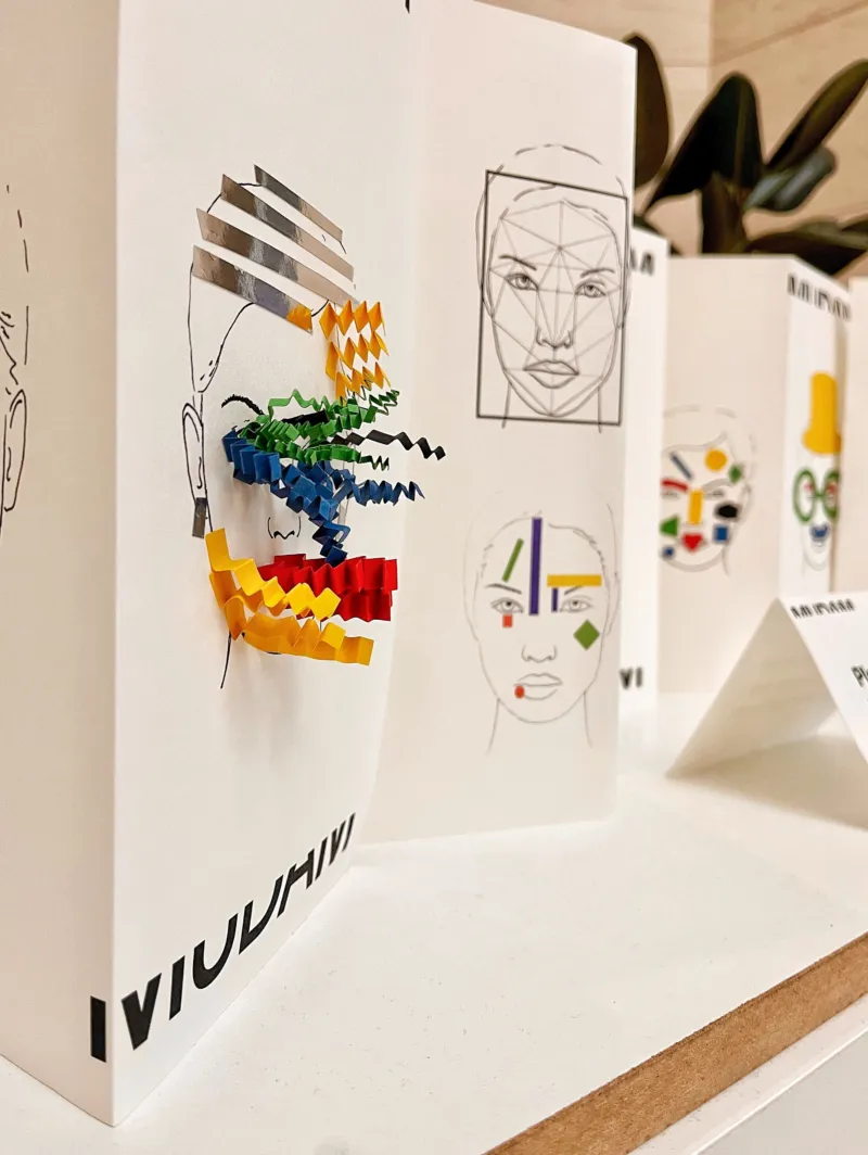 A hand placing papers of different colours, shapes and sizes on the drawing of a face.